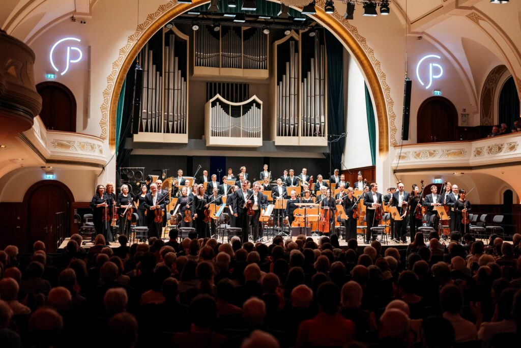 Jenaer Philharmonie auf der Bühne in einem Konzertsaal, mit Publikum im Vordergrund.